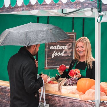 Die Verkäuferin im Marktzelt hält verschiedenes Obst in der Hand. Der Kunde steht vor dem Verkaufszelt an der Theke.