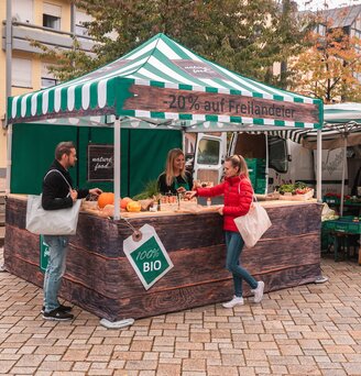 The market tent is fully printed and is used at the weekly market and farmers' market. 2 customers look at the products.