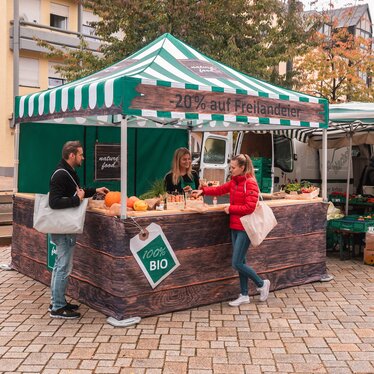 Der Marktstand ist vollflächig bedruckt und kommt auf dem Wochenmarkt und Bauernmarkt zum Einsatz. 2 Kunden sehen sich die Produkte an.