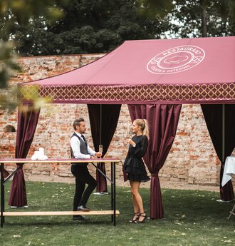 The bordeaux-coloured catering tent stands in the large garden. The woman and the man have a champagne glass in their hands and are standing at a high table.