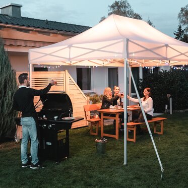 Friends celebrate a barbecue and garden party under the garden tent. The garden tent is festively decorated with fairy lights.