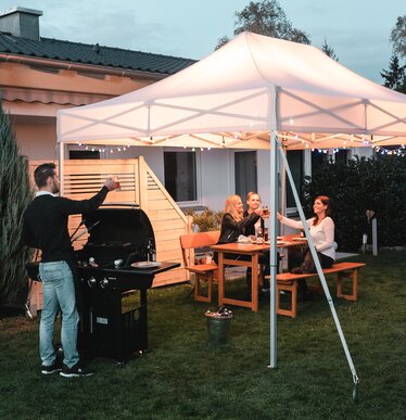 Freunde feiern unter dem Gartenzelt eine Grill- und Gartenparty. Das Gartenzelt ist feierlich dekoriert mit Lichterketten.