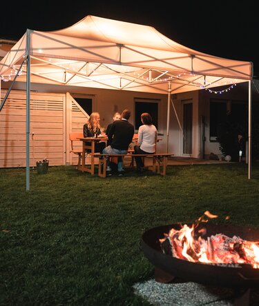 The 4.5x3 m garden tent stands in the garden in the dark. The 4 guests sit under it. There is a fireplace in front of the gazebo.