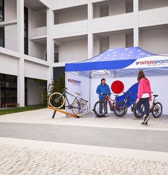 The printed promotion tent from "Intersport" stands on the outdoor area of the fair. Under the 4.5x3 m folding gazebo stands the salesman and his bicycles.