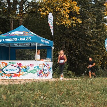 The blue and white event tent serves as a supply station for a marathon. 2 runners are passing the event tent.