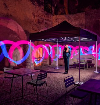 The black folding gazebo stands in a courtyard. Under it stands a young woman. Around the folding tent there are light strips in pink and blue.