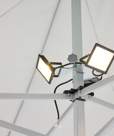 Der LED-Scheinwerfer, bestehend aus 3 Spots, ist am Mittelpfosten des Faltpavillons montiert. Der LED-Scheinwerfer ist an.