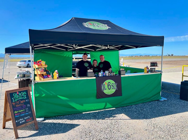 Auf einem Markt steht ein grün-schwarzer, bedruckter Ecotent Faltpavillon Serie E2 der Größe 4,5 x 3 Meter mit Seitenwänden mit Tür und Counter von Ol 6 Six. 